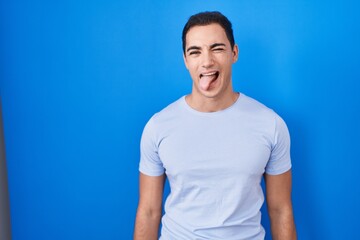 Fototapeta premium Young hispanic man standing over blue background sticking tongue out happy with funny expression. emotion concept.