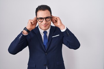 Young hispanic man wearing suit and tie covering ears with fingers with annoyed expression for the noise of loud music. deaf concept.
