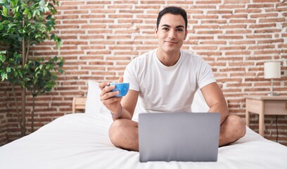 Young hispanic man using laptop drinking coffee at bedroom
