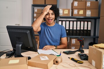 Hispanic man with curly hair working at small business ecommerce surprised with hand on head for...