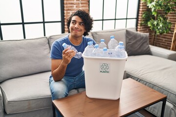 Hispanic man with curly hair holding recycling bin with plastic bottles at home smiling looking to...