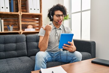 Hispanic man with curly hair doing online session at consultation office pointing thumb up to the side smiling happy with open mouth