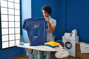 Hispanic man with curly hair ironing holding burned iron shirt at laundry room skeptic and nervous,...