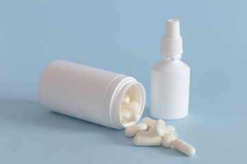 White medicine bottle without a lid lying down with the fallen capsules and a nasal spray next to it on a blue background.