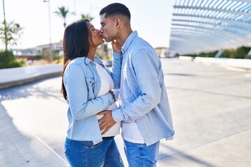 Young latin couple expecting baby touching belly and kissing at street