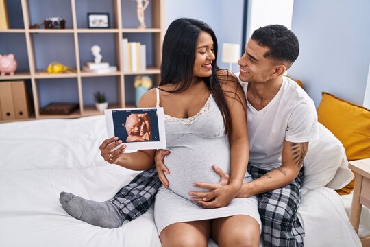 Young latin couple expecting baby hugging each other holding ecography at bedroom