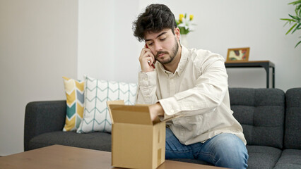 Young hispanic man speaking on the phone unpacking cardboard box at home