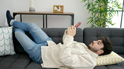 Young hispanic man using smartphone lying on sofa at home