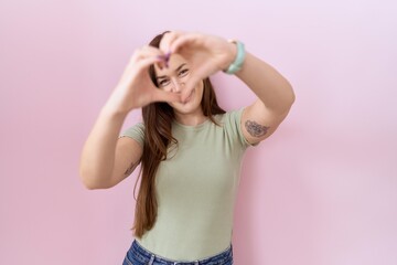Obraz na płótnie Canvas Beautiful brunette woman standing over pink background smiling in love doing heart symbol shape with hands. romantic concept.