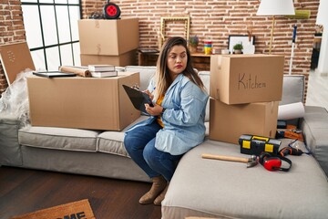Young beautiful plus size woman sitting on sofa holding clipboard at new home