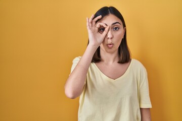 Hispanic girl wearing casual t shirt over yellow background doing ok gesture shocked with surprised face, eye looking through fingers. unbelieving expression.
