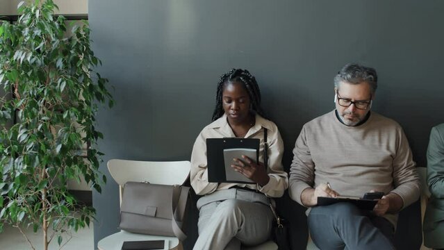 Medium shot of black woman, Caucasian woman and Caucasian man waiting in line for job interview, looking through their CVs, then black woman taking eyes from hers, looking at camera