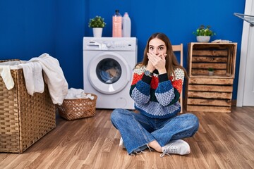 Young hispanic girl doing laundry shocked covering mouth with hands for mistake. secret concept.
