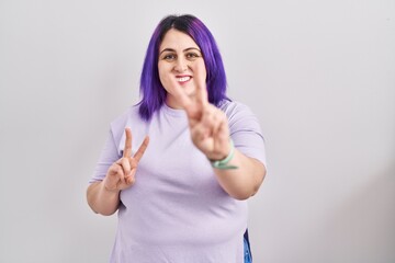 Obraz na płótnie Canvas Plus size woman wit purple hair standing over isolated background smiling looking to the camera showing fingers doing victory sign. number two.