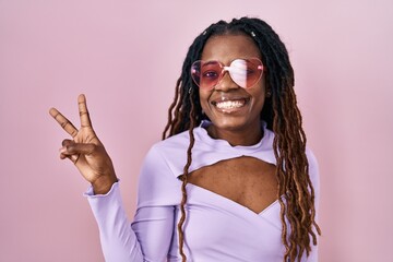 African woman with braided hair standing over pink background smiling with happy face winking at the camera doing victory sign. number two.