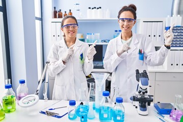 Fototapeta premium Young mother and daughter at scientist laboratory smiling and looking at the camera pointing with two hands and fingers to the side.