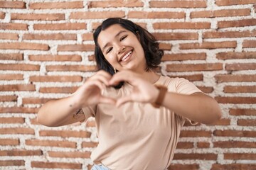 Young hispanic woman standing over bricks wall smiling in love doing heart symbol shape with hands. romantic concept.