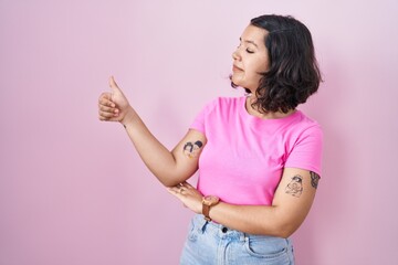 Young hispanic woman standing over pink background looking proud, smiling doing thumbs up gesture to the side