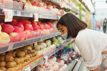 Portrait of healthy young Asian woman with casual clothes is wearing medical face mask when buy fresh organic fruit to make healthy food with new normal lifestyle  and social distancing at supermarket