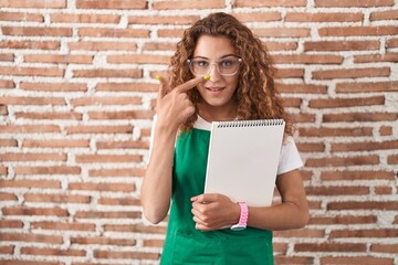 Young caucasian woman holding art notebook pointing with hand finger to face and nose, smiling cheerful. beauty concept