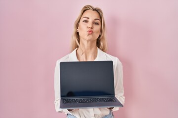 Young caucasian woman holding laptop showing screen looking at the camera blowing a kiss being...