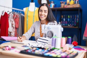Fototapeta premium Young beautiful hispanic woman tailor smiling confident drawing on notebook at clothing factory