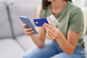 Young beautiful hispanic woman using smartphone and credit card holding pills at home