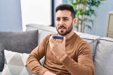 Young hispanic man talking on the smartphone sitting on sofa at home