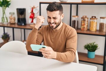 Young hispanic man using smartphone sitting on table at home