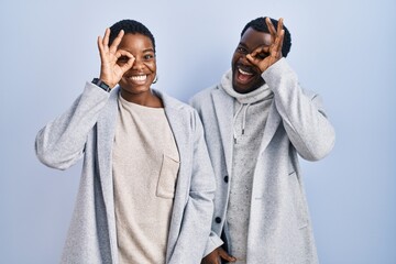 Young african american couple standing over blue background together doing ok gesture with hand smiling, eye looking through fingers with happy face.