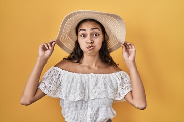 Young brazilian woman wearing summer hat over yellow background puffing cheeks with funny face. mouth inflated with air, catching air.