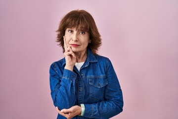 Middle age woman standing over pink background thinking worried about a question, concerned and nervous with hand on chin