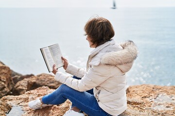 Middle age woman reading book sitting on the rock at seaside