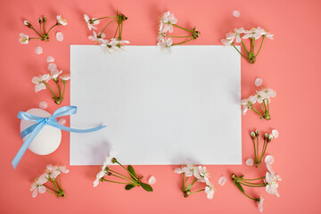 A white sheet of paper, cherry blossoms and an egg with a blue ribbon on a pink background. Easter background, copy space. Flatlay
