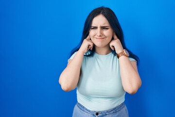 Young modern girl with blue hair standing over blue background covering ears with fingers with annoyed expression for the noise of loud music. deaf concept.