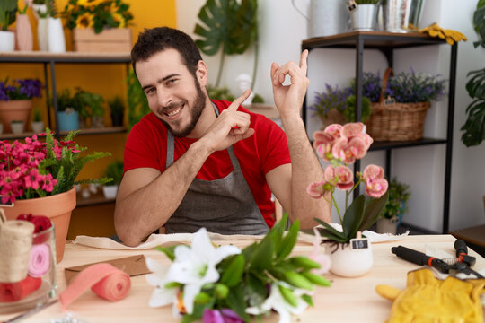 Young hispanic man working at florist shop smiling and looking at the camera pointing with two hands and fingers to the side.