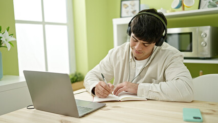 Young hispanic man student using computer taking notes wearing headphones at dinning room