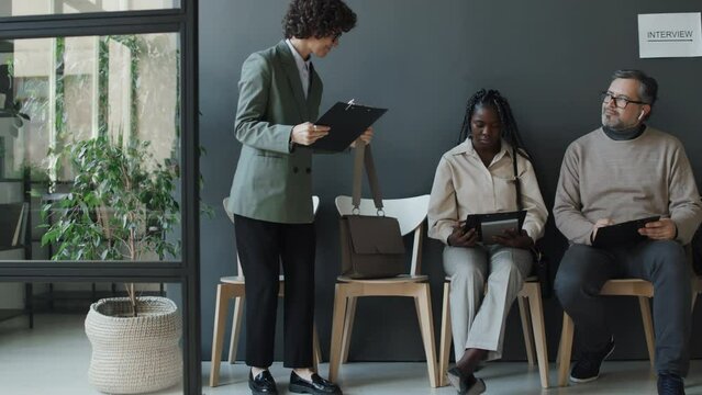 Full shot of Caucasian woman enetring hall and sitting beside black woman who looking at her and Caucasian man, all holding CV, waiting for interview