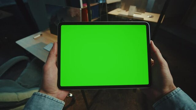 In dark office man holding scrolling a tablet computer with a horizontal green screen. Mock-up display. Device. Apartment