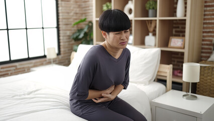 Fototapeta premium Young chinese woman suffering for menstrual pain sitting on bed at bedroom
