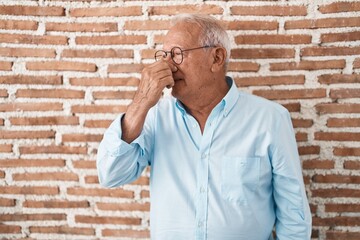 Senior man with grey hair standing over bricks wall smelling something stinky and disgusting, intolerable smell, holding breath with fingers on nose. bad smell