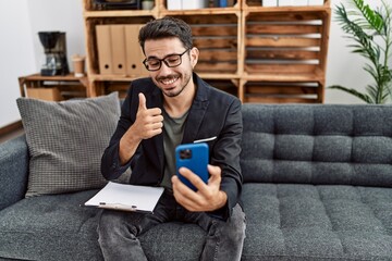 Young hispanic psychologist man doing therapy on video call with smartphone smiling happy and positive, thumb up doing excellent and approval sign