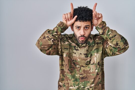 Arab man wearing camouflage army uniform doing funny gesture with finger over head as bull horns