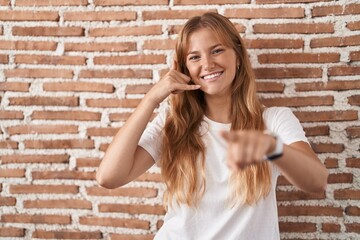Young caucasian woman standing over bricks wall smiling doing talking on the telephone gesture and pointing to you. call me.