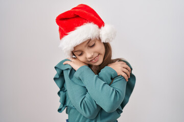 Little caucasian girl wearing christmas hat hugging oneself happy and positive, smiling confident. self love and self care