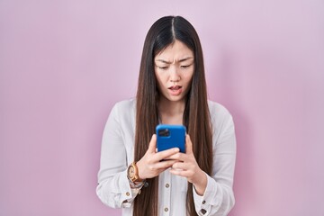 Chinese young woman using smartphone typing message in shock face, looking skeptical and sarcastic, surprised with open mouth