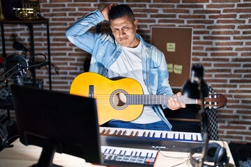 Hispanic young man playing classic guitar at music studio confuse and wondering about question. uncertain with doubt, thinking with hand on head. pensive concept.