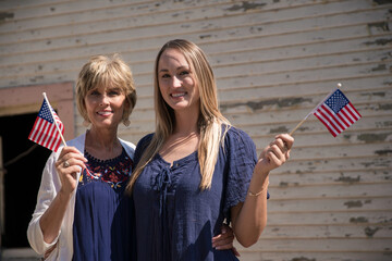 This image shows a mother and daughter posing together, smiling, and waving patriotic American...