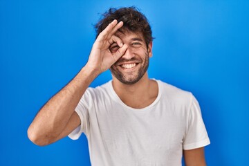 Hispanic young man standing over blue background doing ok gesture with hand smiling, eye looking through fingers with happy face.