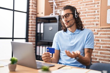 Young hispanic man call center agent holding credit card speaking at office